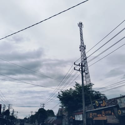 The image presents a street scene in Lada, Indonesia, under an overcast sky filled with grey clouds. A tall, metal lattice telecommunications tower stands prominently on the right side of the frame, reaching high into the sky and equipped with multiple antennas.

The scene is dominated by a dense network of overhead utility lines and poles. Numerous black cables crisscross the sky, supported by both wooden and concrete utility poles that line the street, suggesting extensive electrical and communication infrastructure.

Along the right side of the road, several single-story buildings with corrugated metal roofs are visible, appearing to be local shops or small businesses. Attached to one of these buildings is a large sign displaying the brand "DJARUM D" with an image of cigarettes, marked "Rp 15.000" and "BARU" (new). Below this, multiple banners feature the brand name "RAPTOR," accompanied by text such as "BEBAS MENTO" and further instances of "RP. 15.000."

The street stretches into the distance, with what appears to be a dark-colored vehicle, possibly a truck, visible in the middle ground. Further down the road, blurry figures of people and motorbikes suggest general street activity. Trees and other vegetation are interspersed among the buildings and utility structures. The overall impression is a typical daytime street view in a town, characterized by utilitarian structures and the subtle presence of local commerce, all under diffuse light from the cloudy weather.