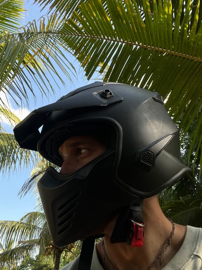 A person wearing a matte black motorcycle helmet with a protective face mask stands outdoors, partially obscured by palm fronds. The sky is blue with scattered clouds. The helmet has a prominent visor and several vents. The person's visible eye, a portion of their neck, and a chain necklace are also visible. The image appears to be taken at ground level, looking up. The location is specified as Kerambitan, Indonesia.