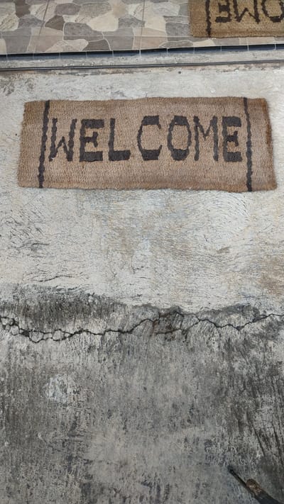 A coir doormat with the word "WELCOME" in dark lettering is placed on a cracked concrete surface. The doormat is light brown with darker brown stripes on the sides and text. Above the doormat, there is a tiled surface with a pattern of interlocking geometric shapes in shades of gray and beige. Another doormat, partially visible to the right and further up, also displays text. The scene appears to be an outdoor entrance. The image is taken from a high angle, looking down at the doormat and surrounding ground.