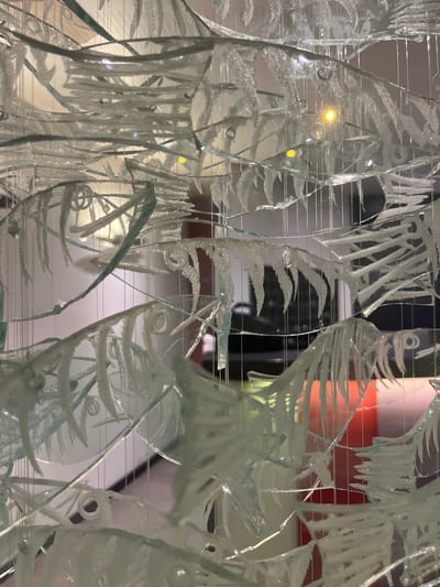 The media file presents a close-up, abstract view of a decorative glass art piece, likely a sculpture or installation. The glass is fragmented and layered, with etched or frosted designs that suggest stylized fish and aquatic foliage. Thin, horizontal wires are visible, interspersed throughout the glass panels, hinting at a suspension system for the artwork. The lighting creates reflections and refractions, adding depth and a shimmering quality to the glass. One bright yellow light source is prominently visible, appearing to be a bulb within or behind the artwork. The background is blurred and indistinct, showing hints of color, including orange and white, and what might be other parts of an interior space. There are no visible people or overt signs of activity, suggesting the image captures a static artwork. The overall impression is one of intricate craftsmanship and artistic interpretation of nature, creating an ethereal and somewhat fragmented aesthetic. Given the location context of Geneva, Switzerland, this could be an exhibit in a museum, gallery, or public space. The time of day and weather are not discernible. No text is visible.