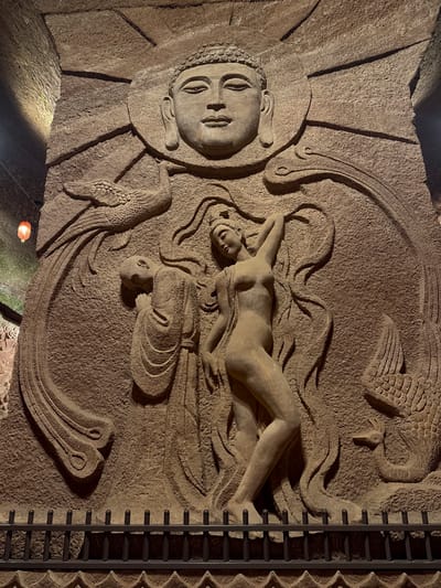 This image depicts a stone relief sculpture, possibly a wall carving, featuring a central reclining nude figure with two other faces visible. The sculpture includes symbolic elements such as waves, sun rays around a face, and stylized birds. The carving style seems ancient or traditional. The scene is illuminated with dim light. A metal railing or frame is visible on the right side of the image. It is known as the Dazu Rock Carvings, located in Shizhong District, Dazu, Chongqing, China. It is listed as a UNESCO World Heritage site.