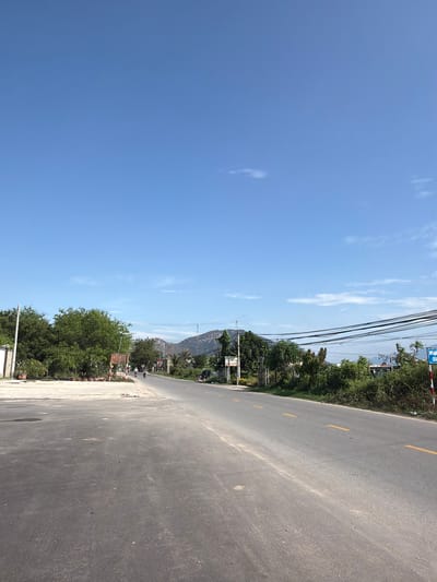 The image displays a wide, paved asphalt road stretching into the distance under a bright, clear blue sky dotted with sparse, wispy white clouds. A yellow dashed line is visible on the road surface, which appears somewhat dusty. Lush, dense green trees and vegetation line both sides of the road, forming a vibrant natural barrier. In the background, rugged mountains or hills rise, partially obscured by the foliage and a network of black power lines strung across utility poles.

On the right side of the road, a blue and white sign is partially visible, displaying Vietnamese text which includes "CÂY XĂNG" (gas station) and "SẠCH" (clean). Another brown sign is present further down the road, nestled among the trees. The scene is bathed in bright daylight, indicating mid-morning or early afternoon. The overall setting suggests a calm, rural or semi-urban landscape in Ninh Chu Ward, Vietnam, with no prominent human activity or vehicles immediately visible, conveying a sense of tranquil roadside scenery.
