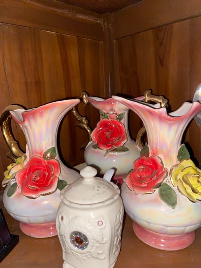 A collection of three decorative vases and a lidded sugar bowl are arranged on a wooden shelf. The vases are made of porcelain with an iridescent finish, displaying shades of pink, yellow, and white. Each vase is adorned with raised rose embellishments in red and yellow, accented with green leaves and gold trim. The handles and spouts of the vases are also gold-colored. In front of the vases sits a white porcelain sugar bowl with a lid and a small spoon. The sugar bowl features intricate embossed designs and is decorated with several colorful gemstones. The background is a wooden cabinet with vertical grain. The image appears to be taken indoors, likely in a domestic setting within Margorejo, Indonesia. There are no people visible in the scene.