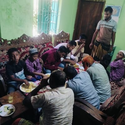 This image captures a communal meal taking place inside a home in Lalmohan, Bangladesh, during the daytime, suggested by the bright light entering through a window.

The scene features a group of approximately ten to twelve males, ranging from young boys to adults. Most are seated on a large, ornately carved wooden sofa with red and black patterned upholstery, or on cushions on the floor. In the foreground, a wooden coffee table holds a glass of water and a small white plate with sliced cucumbers and chilies.

Several large plates, filled with a rice-based dish (possibly biryani or pulao), are distributed among the group, from which individuals are eating directly with their hands. The atmosphere appears relaxed and social, with everyone focused on the meal. Notable individuals include an adult male with a reddish-orange beard and hair, and several boys and men wearing traditional Islamic skullcaps. One adult male, wearing a striped polo shirt and a lungi, stands near a wooden door in the background.

The room features light green painted walls and a large window fitted with decorative metal grates. A calendar or chart displaying anatomical diagrams, including a cross-section of a tooth, hangs on the wall behind the standing man, though any text on it is illegible. The overall setting suggests a lived-in, modest, and culturally typical Bangladeshi household.