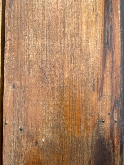 This image depicts a detailed, vertical close-up of an aged wooden surface. The wood exhibits a primary reddish-brown coloration with prominent, dark vertical grain patterns. A distinct, darker, almost black, vertical band of discoloration or natural wood variation is visible along the rightmost portion of the primary wooden panel. A narrower, darker vertical strip runs along the far left edge, indicating a seam or separate component. The surface possesses a worn, textured appearance with fine vertical striations consistent with wood grain. Several small, dark, roughly circular indentations are present, consistent with nail holes or small knots, with specific examples in the lower left and lower right quadrants. A small, irregular white speck is visible in the lower-middle left. No individuals, animate subjects, or discernible environmental context beyond the wooden surface are present. The scene is located in the city of Chascomús, Argentina.