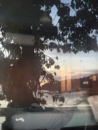 The image depicts an exterior view captured from an interior vantage point in Koh Tao, Thailand, during either sunrise or sunset. In the immediate foreground, a bed is visible, covered with light grey sheets and a dark garment draped upon it. A partial white object, likely furniture, is present on the right edge. The midground is dominated by the dark silhouette of a large tree with dense foliage. A white outdoor air conditioning unit and a light-colored ceiling fan are visibly attached within the tree's canopy. Beyond the tree, a calm body of water extends towards a distant landmass on the horizon. The sky displays hues of orange, pink, and light blue, indicative of low sun angles. The distant landmass features vegetation, multiple square-shaped structures, and several small piers or jetties extending into the water. Reflections of the sky and distant elements are discernible on the water's surface. No individuals are present in the scene.