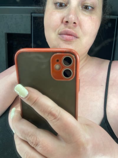The image is a close-up selfie taken by a person holding a smartphone in their right hand, in what appears to be a domestic setting. The person's face is visible, with their eyes looking towards the phone's camera. The phone has an orange-rimmed case with a dual camera lens. The person's left arm and shoulder are also visible, with a black strap of a garment. The background is dark and out of focus, suggesting an indoor environment. There is no information about the location in the city of Rozkvit, Ukraine.