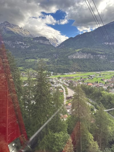 A panoramic view of the Swiss Alps and the town of Meiringen, Switzerland, is captured from a cable car. The rugged, mist-shrouded peaks of the Alps dominate the upper portion of the frame, with dense green forests blanketing their lower slopes. Below, the picturesque town of Meiringen nestles in the valley, characterized by its charming architecture with red-tiled roofs, nestled amidst lush green fields and winding roads. A river snakes through the landscape, and a pedestrian bridge is visible. In the foreground, tall evergreen trees obscure parts of the view, and a red reflective surface on the left suggests the cable car window. The sky is a dramatic mix of dark, stormy clouds and patches of bright blue, hinting at changing weather conditions.