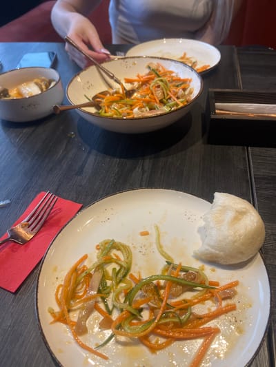 The image captures a close-up view of a dining table with partially eaten food. In the foreground, a white plate holds remnants of a salad composed of julienned carrots and cucumbers, along with some protein, possibly meat or tofu. A soft, bun-like food item is visible on the right side of the plate, with a bite taken out of it. A silver fork rests on a red napkin to the left of the plate. In the background, two bowls contain more food, with one appearing to hold a soup-like dish with mushrooms and egg. A person's hands, with painted nails, are visible reaching towards one of the bowls. The table surface is dark and appears to be made of wood. The overall setting suggests a meal in progress or recently concluded in a restaurant. There is no identifiable information about the location being Angarsk, Russia within the image.