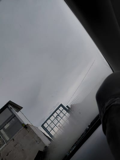 This image, taken in Zagore, Bulgaria, captures a view upward through what appears to be a vehicle's windshield on an overcast day. The dominant feature is a uniform, light-gray, cloudy sky, filling most of the frame and suggesting daytime without direct sunlight.
Parts of the vehicle's interior are visible in the upper left and top center, including a dark, curved element possibly from the dashboard or rearview mirror area, and a dark frame line consistent with a car's roofline or A-pillar. A prominent, long, thin dark line, likely a crack in the windshield or an external wire, stretches diagonally across the sky in the upper-mid section.
In the middle-right, a large, grid-like structure, possibly scaffolding, an antenna array, or part of a building's roof, is reflected or seen through the glass, displaying a metallic or light-colored appearance.
Towards the lower right, a partial view of a building made of light-colored concrete or stone is visible. This building features large glass windows or a facade. A security camera is mounted on its exterior, and below it, a small, unreadable yellow-green sticker or sign is present.
No people are visible, and no specific activity is taking place. The scene conveys a sense of quiet observation from within a vehicle, under dull, cloudy weather conditions.