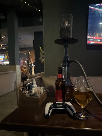 An overhead shot, viewed from an eye-level perspective looking down onto a wooden table, captures a dimly lit indoor setting, likely a bar or lounge in the evening.
In the foreground, a prominent black hookah with a mesh top and dark stem stands on the table. A flexible black hose extends from the hookah. Beside it are several brown glass bottles, one of which appears to have a red label. A clear wine glass containing an amber-colored liquid, possibly beer, is tilted on its side near the bottles. To the right of the glass, a white and black gaming controller, resembling a PlayStation DualSense, rests on the table. A small, dark wooden caddy filled with various small containers, possibly hand sanitizers or other amenities, is also visible on the table to the right.
In the background, a large television screen displays what appears to be a sports match, with the word "BENFICA" clearly visible, suggesting a soccer game involving the Portuguese club S.L. Benfica. Below the TV and extending further into the background, other parts of the lounge are visible, characterized by soft, warm string lights, blurred figures of people, and comfortable seating, creating a relaxed and social atmosphere. The overall scene suggests a casual entertainment space where patrons can enjoy drinks, hookah, gaming, and sports.