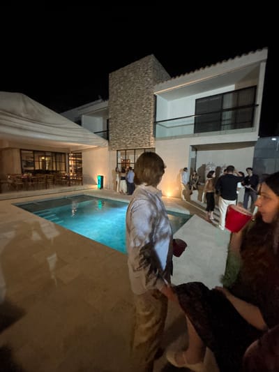 A nighttime scene in Santa Cruz Tehuixpango, Mexico. A group of people are gathered around a pool. A two-story building is visible, with windows and a textured facade. A covered walkway with tables and chairs is adjacent to the pool. People are interacting near the pool and building. One person is closer to the camera, wearing a striped shirt and looking toward the pool. Another person is partially visible in the foreground. A red cup is present among the group of people. The pool is lit with blue light.