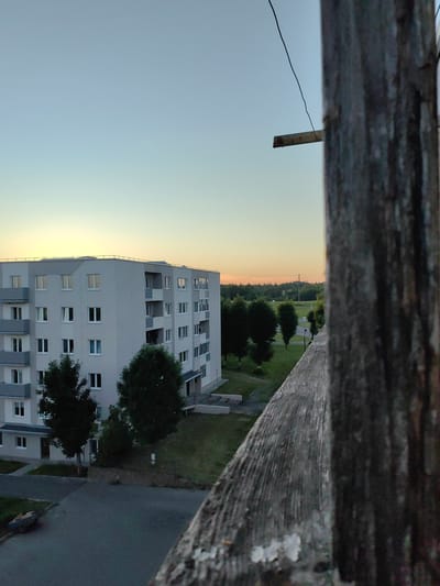 The image captures an exterior view from an elevated perspective, likely a balcony or window, overlooking a residential area in Myadzyel, Belarus. In the foreground, a weathered, textured wooden railing or post dominates the right side, its rough surface and vertical grain prominently displayed, with some white peeling paint evident. Beyond this immediate structure, a modern, multi-story apartment building is visible on the left. The building is light gray with darker gray accents around balconies and window frames, featuring numerous windows and balconies. Trees with dark green foliage are situated in front of and around the building, with a neatly mown lawn. The background reveals a horizon with a line of trees and a pale, gradient sky transitioning from light blue at the top to a soft yellow-orange glow near the horizon, suggesting either sunrise or sunset. A thin, dark wire is visible extending across the sky, attached to a small metal protrusion on the wooden post. The overall scene is quiet and depicts a typical residential landscape. No people are visible in the frame.