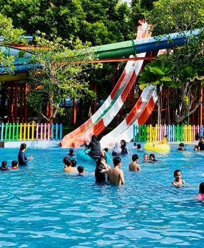 In Pantai Cermin Kiri, Indonesia, an outdoor swimming pool is filled with people enjoying a sunny day at a water park. Several water slides, featuring red, white, and green sections, are visible in the background, with lush green trees surrounding the area. A vibrant, multi-colored fence encloses part of the pool. In the water, numerous individuals of varying ages are engaged in activities, including swimming, playing, and lounging. Some are using inflatable tubes and rings. A few people are standing at the edge of the pool, while others are wading or swimming deeper into the water. The atmosphere appears lively and recreational.