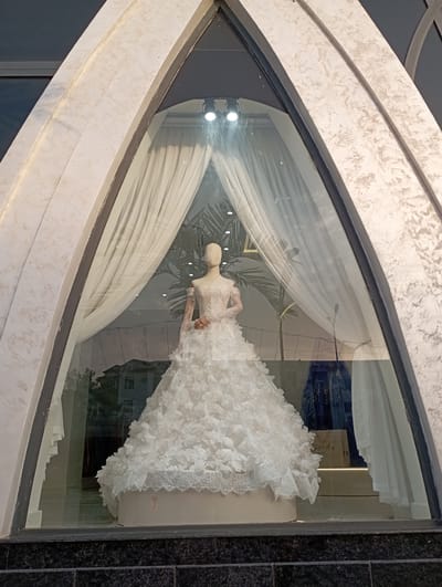 The image shows a bridal shop window display featuring a white wedding dress on a mannequin. The dress is elaborate, with a full skirt adorned with floral appliques and lace trim. The mannequin is positioned in the center of the display, facing towards the right. Behind the mannequin, white drapes are arranged to create a soft, elegant backdrop. Through the window, reflections of the surrounding environment are visible, including buildings and trees, suggesting the display is located on a street or in a public area. The window itself is framed by a textured, light-colored material. The overall scene suggests a formal or celebratory occasion, likely a wedding. The image appears to have been taken during the daytime, with ample natural light illuminating the display.