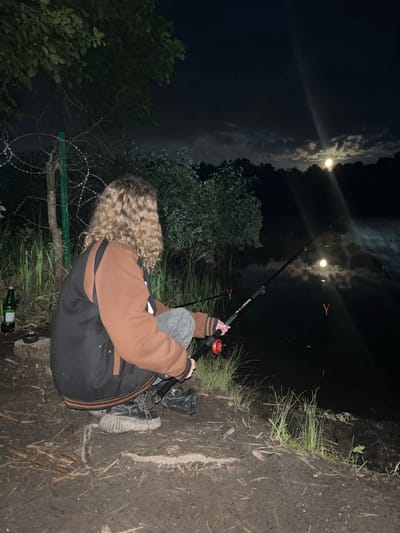 A person is fishing at night in an area near water. The person is holding a fishing rod, and there are fishing accessories present. A bottle is visible on the ground near the person. The environment includes grass, trees, and a barbed wire fence. The moon is visible in the night sky, with its reflection in the water. The location is Krasnyi Jar, Russia.
