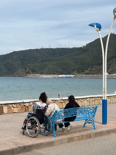 The image shows a seaside scene in M'Diq, Morocco. A blue ornamental bench sits on a paved walkway next to a low stone wall that separates the walkway from the water. Two individuals are on the bench: one seated upright and one leaning forward. A wheelchair is adjacent to the bench, and a woman seated in it is also leaning forward. The scene suggests observation of the water or surroundings. The background reveals a mountain covered in vegetation, a breakwater constructed of large rocks, and a blue sky with light clouds. A distinctive white streetlamp with curved arms and a blue cap stands beside the bench.