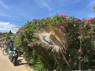 The image depicts a sunny outdoor scene with a large straw hat hanging on a hedge of pink and purple flowers. The hat has blue embroidered lettering that appears to say "Jacqueline". In the background, several people are visible, some walking and one on a bicycle. A black scooter with flower decals is parked in the foreground. Signs are visible on the hedge, one reading "Cabinet Medical" and another "WC PUBLIC", suggesting the location is a public area. The environment is lush and green, with a clear blue sky and some clouds. The scene is likely in a Mediterranean setting, given the vegetation and the overall ambiance, and possibly within Hyères, France, a city known for its coastal beauty and flora.