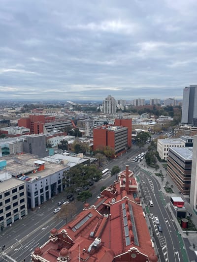 The image provides an elevated, oblique view of a city street scene in Melbourne, Australia, under an overcast sky. The foreground features the distinctive red-tiled roof of an older, ornate building with domed sections and skylights. Adjacent to this, a modern, multi-story building with a pale facade and prominent windows is visible.

A street runs diagonally through the mid-ground, with traffic including cars, a bus, and a white van. Road markings indicate multiple lanes, including designated bicycle lanes in green. Several parked cars and pedestrians are also discernible along the street and sidewalks.

Further into the background, a dense urban landscape unfolds with various buildings of differing architectural styles and heights. Dominating the mid-to-far background are several tall, modern residential or office towers, some with lighter facades and others appearing to be glass-fronted. Red brick buildings, some with multiple stories, are interspersed throughout the cityscape, contributing to the varied urban fabric. The overall impression is one of a busy, developed city center with a mix of historic and contemporary structures. The cloudy weather casts a muted light over the scene.