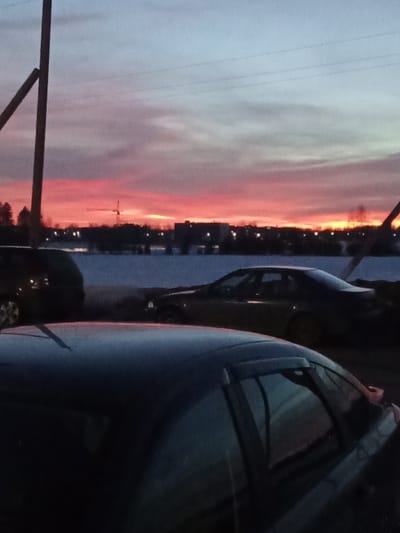 The image displays an outdoor twilight scene in Дашкаўскі сельскі Савет, Belarus, characterized by a vibrant sunset or sunrise. The sky dominates the upper two-thirds of the frame, exhibiting a strong gradient from intense orange and red at the low horizon, transitioning through shades of pink and purple, to light blue at the top. Wispy clouds are present, and multiple thin power lines stretch across the upper sky.
In the midground, a snow-covered landscape extends horizontally, featuring a dark silhouette of trees and buildings along the distant horizon. A construction crane is visible as a dark silhouette against the brightly colored sky on the left side of the horizon. Several small, distant lights are visible, indicating an illuminated area beyond the snow.
The immediate foreground is occupied by three vehicles. The dark-colored roof and antenna of a car are prominently visible at the bottom of the frame, reflecting the sky's colors. Behind this car, to the left, a dark sport utility vehicle (SUV) or hatchback is partially visible, showing its rear wheel and part of its body. To the right of the SUV, a dark sedan is parked, showing its side profile facing right. No individuals are visible in the scene. The overall environment suggests a cold, potentially winter, evening or morning.