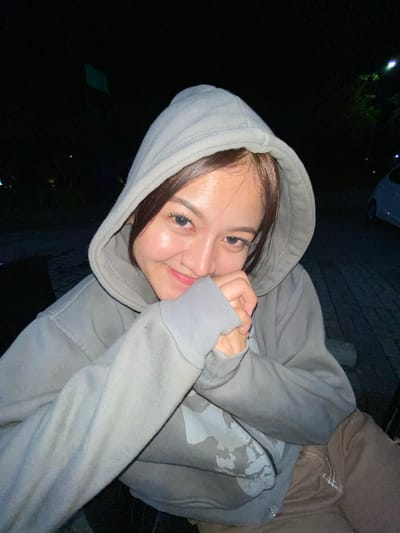 A young woman is captured in a close-up, low-light portrait, wearing a grey hooded sweatshirt. She is looking towards the camera with a shy smile, her hands clasped in front of her face, partially obscuring her mouth. Her hair is dark brown and frames her face. The background is dark, suggesting an outdoor setting at night, possibly in the city of Mekar Bakti, Indonesia. A paved area is visible behind her, and faint lights and greenery can be seen in the distance, along with the outline of a vehicle. The lighting in the image is warm and focused on the woman's face, creating a soft glow.