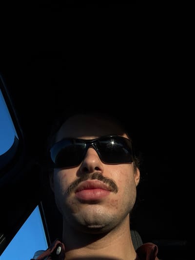 A male individual is depicted in a close-up, head-and-shoulders portrait, facing directly towards the camera within a vehicle in Lodi Vecchio, Italy. He wears dark, wraparound-style sunglasses and has a prominent dark mustache. Strong, direct sunlight illuminates the lower portion of his face, including his lips, chin, and mustache, while his forehead and the upper part of the vehicle's interior are in deep shadow. A circular, translucent patch is visible on the left side of his chin. He is wearing a reddish-brown collared shirt, partially visible at the bottom of the frame. A segment of clear blue sky is visible through a vehicle window on the left, contrasting with the dark interior.