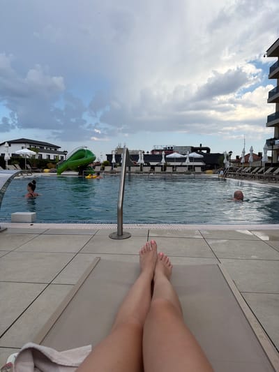 This media file captures a resort pool scene in Hisarya, Bulgaria. The perspective is from someone relaxing on a lounge chair, with their legs and feet, painted with light pink nail polish, visible in the foreground. The pool itself is a prominent feature, with clear blue water reflecting the cloudy sky.
In the pool, a man with a bald head is swimming, his upper body visible above the water. Further into the pool, a woman with her hair tied up is also swimming. A bright green, dinosaur-shaped water slide is visible on the left side of the pool, adding a playful element to the scene.
Surrounding the pool are lounge chairs with white umbrellas, suggesting a leisurely atmosphere. Buildings, appearing to be part of the resort, are visible in the background, featuring dark facades and multi-story structures with visible windows. Some of the buildings have visible text, though it is not clearly legible.
The sky is filled with dramatic, gray and white clouds, indicating an overcast or potentially rainy day. The time of day appears to be late afternoon or early evening, given the soft lighting. The overall mood of the image is relaxed and vacation-oriented, with the presence of the pool, lounge chairs, and people enjoying the water. The presence of the slide suggests it might be a family-friendly resort.