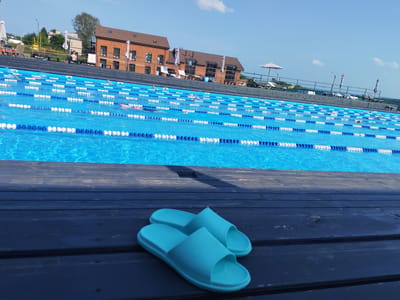 In Pirogovo, Russia, a bright blue swimming pool with clear water is divided into lanes by white and blue buoys. Several swimmers are visible in the pool, some actively swimming and others appearing to float. In the foreground, a pair of light blue shower sandals rests on dark wooden planks. Behind the pool, a multi-story brick building with large windows and a sloped roof is visible, along with lounge chairs and umbrellas. The sky is clear and blue with a few scattered white clouds.