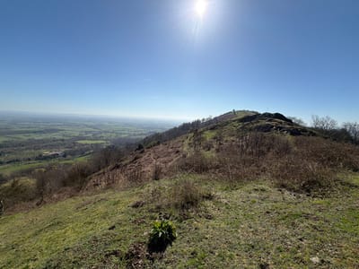 The image displays an elevated landscape scene within the Malvern Hills, United Kingdom, on a clear, sunny day with the sun visible in the upper-left quadrant. The foreground consists of a gently sloping area covered in patchy green grass and brown, dry vegetation, with a small cluster of bright green foliage centrally located at the bottom. The midground features a prominent hillside or ridge, partially covered with rocky outcrops near its crest, and slopes characterized by sparse deciduous trees, some bare, intermingled with green grass and brown undergrowth. A dirt path or trail is discernible traversing the hillside on the left, leading towards a faintly visible small marker or structure on a more distant, higher point of the ridge. The background extends into a vast, panoramic view of a valley and plains below, presenting a mosaic of green agricultural fields, darker wooded areas, and indistinct distant structures, all receding towards a hazy horizon. No human subjects, animals, or explicit actions are depicted.