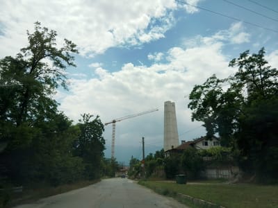 This media file captures a street view in Momin Prohod, Bulgaria, under a cloudy sky. The scene features a road leading towards a construction site, characterized by a tall, white, tower-like structure and a large yellow crane. Dense green trees line both sides of the road and the surrounding area, creating a naturalistic contrast with the industrial elements.
In the foreground, the asphalt road shows signs of wear and tear, with visible cracks and patches. To the right of the road, a grassy area is visible, with a green trash bin and part of a building with a tiled roof. Further down the road, two cars are driving away from the viewer. A traffic sign displaying "50" is present on the right side of the road.
The sky is filled with white, fluffy clouds against a bright blue backdrop, suggesting it's daytime. The overall atmosphere appears to be peaceful, with a juxtaposition of nature and ongoing construction. There are no visible people in the frame, but the presence of cars and the construction site indicate human activity. The composition suggests the photo was taken from inside a vehicle moving along the road.