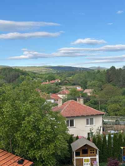 An elevated view of Pchelin, Bulgaria shows a cluster of houses with red tile roofs surrounded by lush green trees. A building with white walls and a red tile roof is in the foreground to the right, adjacent to a wooden playhouse. Overhead power lines are visible. In the distance, hills stretch towards the horizon under a blue sky streaked with white clouds. The weather is sunny. No people are visible.