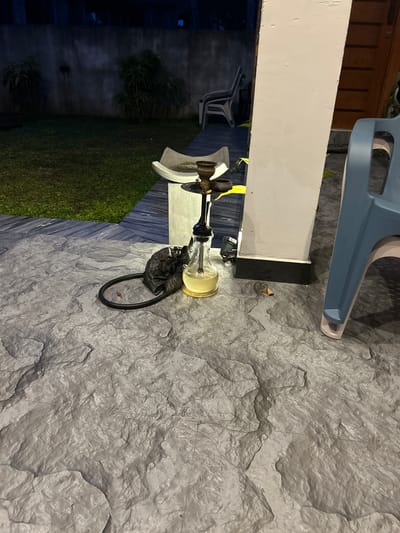 The image depicts an outdoor patio area at night. A hookah pipe, filled with yellow liquid, sits on the textured gray stone floor. A black trash bag and a black hose are next to the hookah. In the background, a grassy yard is visible, bordered by a concrete wall. Two chairs are also present in the scene. The setting appears to be residential, potentially in Kadewatta, Sri Lanka, given the context. There are no people or animals visible in the frame. The lighting suggests it is nighttime.