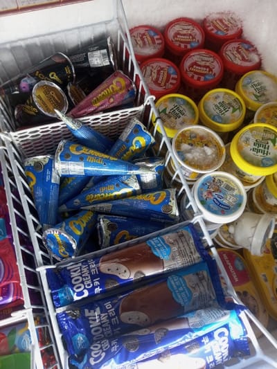 This image displays the interior of a commercial freezer, likely within a retail establishment in Naung Bo Oe, Myanmar. The freezer contains various packaged frozen desserts, primarily organized within white wire baskets. In the center, a large quantity of "Cornetto Midi" brand ice cream cones in blue packaging, featuring Oreo cookie imagery, are loosely stacked. Above these, several "Cornetto" cones in dark brown and pink packaging are visible. To the right, multiple round ice cream tubs are arranged. These include several red-lidded tubs labeled "Cheesecake 2 Senses," numerous yellow-lidded tubs branded "Hula Hula," and several white-lidded tubs, one clearly marked "Happy Cow." Ice crystals are visible on the lids of many tubs. At the bottom of the frame, several individually wrapped "Cookie Creamy" ice cream bars in blue packaging, displaying Korean text, are stacked. The interior walls of the freezer exhibit a noticeable accumulation of frost. No individuals are present, and no actions or interactions are depicted.