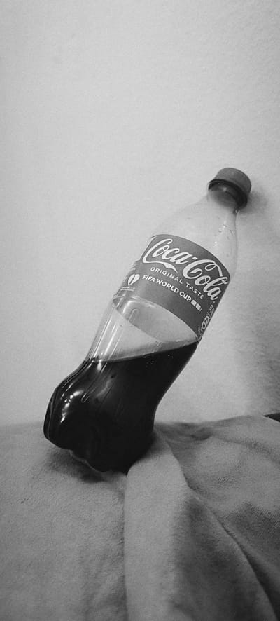 A black and white, close-up, eye-level, medium shot shows a partially filled plastic bottle of Coca-Cola, lying on its side on a textured, light-colored surface, possibly fabric. The bottle is angled upwards, with its cap pointing towards the upper right of the frame. The Coca-Cola logo, in its distinctive red and white script, is visible on the label, which also indicates "ORIGINAL TASTE" and "FIFA WORLD CUP 2026". There is a small, green emblem with a number "0" on the label, and text that reads "ABGEFÜLLT IN DEUTSCHLAND". The liquid inside the bottle is dark brown and opaque. The background is a plain, textured white wall. No people or animals are visible in the scene. The lighting is soft, creating subtle shadows and highlights on the bottle and the surface it rests on.