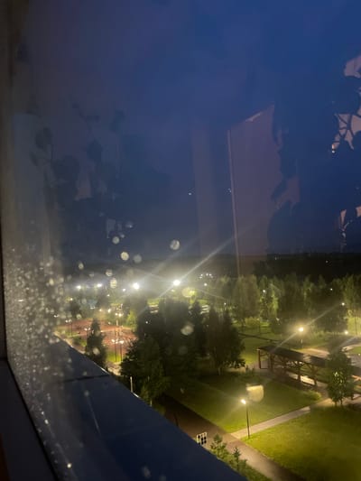 This image, taken at night from a window in Laishevo, Russia, shows a park area with scattered streetlights illuminating green grass and trees. The view is partially obscured by reflections on the glass, including what appear to be leaves and a dark silhouette of a person. In the foreground, the windowpane itself is covered in water droplets. The park features pathways, a playground, and a covered wooden structure. The sky is dark blue.