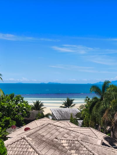This is an elevated, eye-level, medium shot of a tropical beach scene. The foreground is dominated by the tiled roofs of several buildings, with two red satellite dishes visible on one roof. Lush green foliage and palm trees are scattered throughout the middle ground, partially obscuring other buildings and more tiled roofs. The background features a bright, sandy beach that slopes into a calm, turquoise ocean. In the distance, a hazy horizon is punctuated by several mountainous islands. The sky is a vibrant, cloudless blue. The scene suggests a warm, sunny day in a coastal resort area, likely in Baan Thong Po, Thailand, given the tropical vegetation and architecture. There are no people or animals visible in the image.