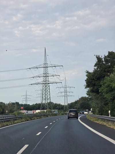 The image shows a multi-lane highway in Raunheim, Germany, with high-voltage power lines crossing overhead. In the foreground, a dark-colored SUV is driving in the right lane, followed by other vehicles. The road is asphalt with clear lane markings. On the left side of the highway, there is a barrier and vegetation, with several large power pylons visible in the distance. On the right side, trees and more vegetation line the road. The sky is overcast.