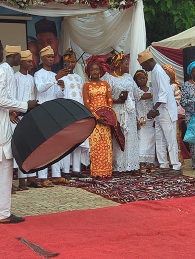 In Kabala, Nigeria, a group of individuals participates in what appears to be a formal event, likely a traditional wedding ceremony, indicated by the 'IYAWO ABIODUN' fan. The scene unfolds on a stage or raised platform covered with a patterned rug and a red carpet.
On the far left, a person in white traditional attire, visible from the torso down, holds a large black octagonal photography softbox with an orange interior, angled towards the central figures. To their right, four young men in white traditional garments and matching light-colored caps are present. One of these men, wearing a darker patterned cap, holds a smartphone up, seemingly capturing a photograph or video.
The central figure is a woman in a vibrant orange and yellow patterned dress and a matching headwrap (gele). She holds a red and gold fan inscribed with "IYAWO ABIODUN" (meaning "Wife Abiodun" in Yoruba). To her immediate right, another woman in a light blue and white lace dress with a gold headwrap holds a dark clutch bag and looks towards her right. A young girl in white lace and a patterned headwrap stands partially visible behind this woman.
Further to the right, a man in white traditional attire and a light-colored cap looks down at a small, ornate golden vessel held by a woman in white lace, whose face is mostly obscured. The man holds a dark smartphone in his left hand. Another woman in a blue and brown patterned dress and blue headwrap stands on the far right.
The background features white and red drapery, with red and white floral arrangements along the top. A large poster depicts a man in traditional Nigerian attire, with another person partially visible behind him. The ground beyond the red carpet appears to be paved with light-colored interlocking stones. The participants are wearing a mix of sandals and closed shoes.