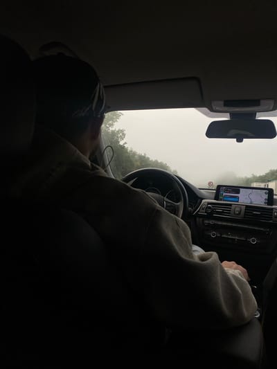 The image depicts the interior of a car. The vehicle is in the vicinity of Beausemblant, France, as indicated by the navigation system on the dashboard display. A person is seated in the passenger seat, leaning back. The car is moving, and the conditions outside are foggy.