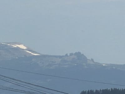 The image shows a distant view of a mountain range under a hazy, light blue sky. Snow is visible on the upper slopes of the mountains, suggesting a cool or mountainous climate. In the foreground, several dark power lines stretch horizontally across the frame, obscuring the lower portion of the mountainous landscape. The overall impression is one of a remote, natural setting, possibly viewed from a distance, with man-made infrastructure interrupting the otherwise pristine scenery. The specific location in Tashtagol, Russia, is not visually discernible from the image itself, as there are no landmarks or identifying features that point to this particular city.