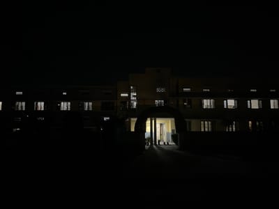 A nighttime, exterior, low-angle shot captures a dimly lit building in Peshawar City Tehsil, Pakistan. The building, possibly a hostel or institutional structure, features a prominent arched entranceway that is illuminated, revealing potted plants and open doorways. Windows on the upper floors are lit from within, casting a warm glow against the dark facade. The surrounding area is cloaked in darkness, with only faint outlines of trees or bushes visible. The image has a slightly grainy quality, indicative of low-light photography.