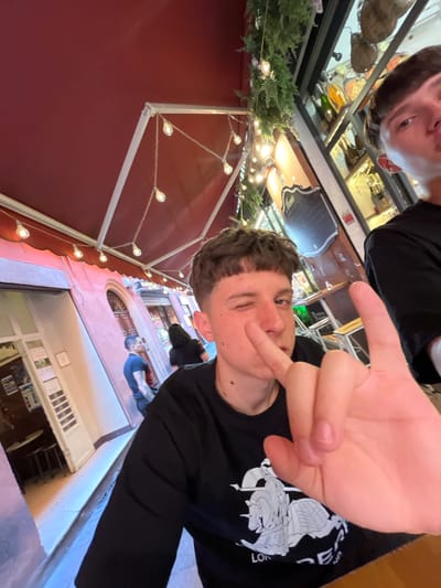 The image is taken in Bologna, Italy. It depicts a selfie taken by a young man with light brown hair, wearing a black long-sleeved shirt with a graphic design on it. He is winking and making a hand gesture with his right hand close to the camera. Another person is partially visible in the upper part of the frame. The scene is in an outdoor restaurant or cafe setting. Behind the subject, the architecture of Bologna is visible. A red awning adorned with lights is above, and beyond that, there is a view down a narrow street lined with buildings, with other people visible in the distance. The image is slightly rotated.