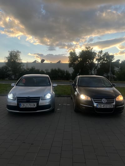 The image captures two Volkswagen cars parked on a paved lot. The car on the left is silver and the car on the right is black. Both cars have their headlights illuminated. The license plate of the silver car reads "CB 2170 OK" and the license plate of the black car reads "CB 4931 CE". In the background, there are trees and a large building with arched structures. The sky is filled with dramatic clouds, illuminated by the setting sun. The scene appears to be in Elin Pelin, Bulgaria, based on the license plates.