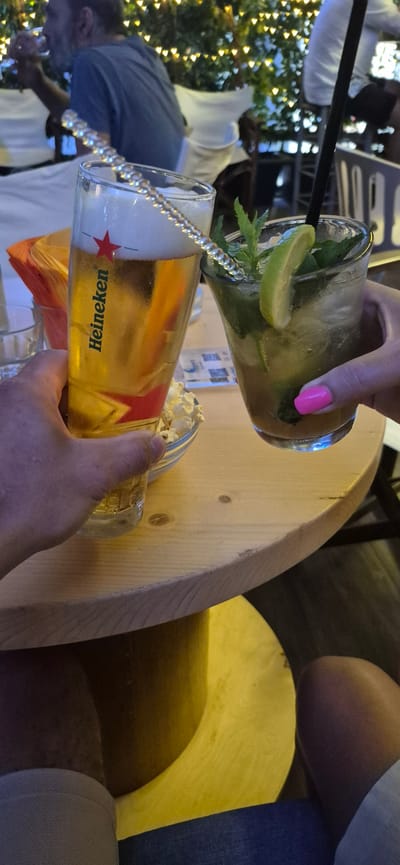 Two people are holding drinks at a table in Paleo Faliro, Greece. One person is holding a glass of Heineken beer with a sparkly straw. The other person is holding a cocktail with mint and lime, and has bright pink nail polish. In the background, there are other people seated and decorative lights. A small bowl of popcorn is also visible on the table. The setting appears to be an outdoor cafe or restaurant with a wooden table.
