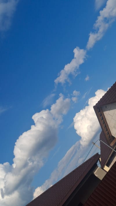 A low-angle outdoor photograph shows a bright blue sky with scattered white clouds. The roofs of two buildings, likely houses, are visible in the foreground and midground, angled diagonally across the frame from bottom left to top right. The roofs are covered in dark brown shingles and appear to be under construction or renovation, with exposed wooden beams and concrete blocks visible on the rightmost structure. The scene is set in Stowbtsy, Belarus. There are no people or animals present in the image.