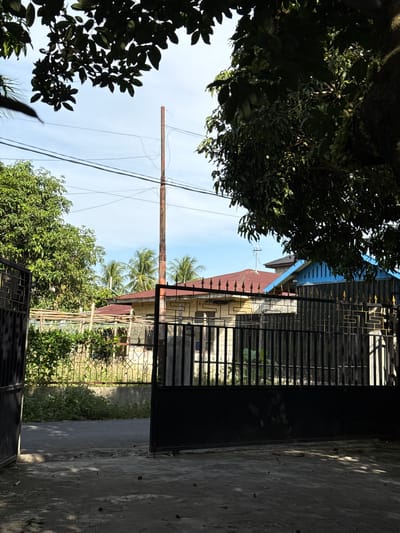 The image depicts an outdoor scene with a clear sky and a few scattered clouds. In the foreground, a dark, open gate leads to a pathway. To the left and right of the pathway, there are areas of rough concrete, with some scattered debris. Beyond the gate, a paved road is visible, bordered by a fence and dense green foliage.

In the middle ground, several houses are visible. One house has a reddish-brown roof and light-colored walls. Another adjacent structure features a blue facade with vertical slats and a reddish-brown roof. A tall, reddish-brown pole stands prominently in the background, with power lines extending horizontally across the sky. Palm trees are scattered behind the houses, suggesting a tropical or subtropical environment. Lush green trees frame the top and right sides of the image, casting shadows on the foreground. The overall scene suggests a residential area. The location is identified as Julicotmeurak, Indonesia.