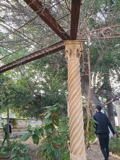 The image is taken from a low angle, looking up at a large, spiraled, white column. The column is part of an elaborate, rusted metal trellis structure, covered in green vines. There are trees growing in the background, with the sun shining through the leaves. There is a person walking away from the camera in the top right corner, wearing a black jacket and black pants. They are walking on a stone pathway. The column is located in the Jardin d’Essai, a botanical garden in Oran, Algeria. The garden is located at 35.705° N, 0.649° W. The garden is a popular place for locals to relax and enjoy the outdoors. There is a second person in the bottom right corner of the image, wearing a black jacket and black pants. This person is walking towards the left edge of the frame. The scene is peaceful and serene. The garden is a welcome escape from the bustling city.