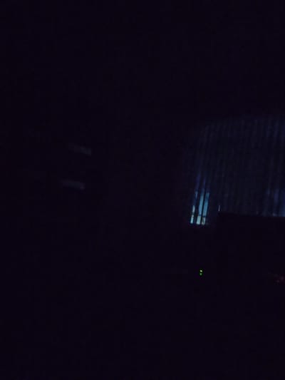 The image depicts a dark interior scene, likely at night, with minimal illumination. On the right side of the frame, a striped pattern, possibly from blinds or a textured surface, is visible, with faint blue light filtering through. Below this, a dark rectangular object is present, with a small green light emitting from its surface. The rest of the image is predominantly black, suggesting a poorly lit environment. Without further context or visual cues, it is impossible to determine the specific location within Baranavichy, Belarus, or identify any people, subjects, objects, or actions.