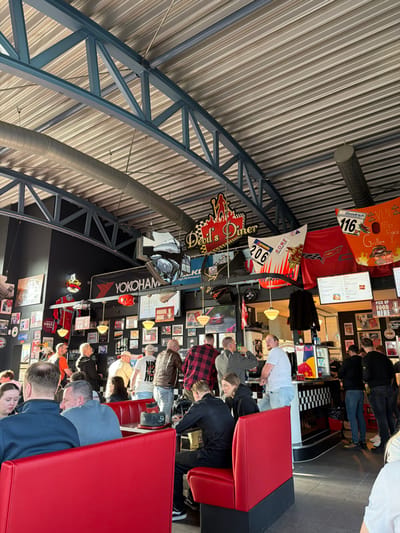 The scene depicts the interior of the "Devil's Diner" in Meuspath, Germany. The establishment features a high, corrugated metal ceiling supported by exposed blue structural trusses, with cylindrical ventilation ducts running horizontally. Walls are dark grey or black and extensively covered with framed photographs, posters, and racing memorabilia. Prominent overhead signage includes a red neon "Devil's Diner" sign, a "YOKOHAMA" banner, and various racing flags/banners, one marked "206" and another "Goodrich VLN 116". Automotive components, including what appears to be a vehicle hood or fender, are suspended from the ceiling structure.
Seating consists of red booths with black bases, several of which are occupied. In the foreground-left, two males and one female are seated at a red booth table. A bald male in a grey shirt sits facing right, while a male in a dark blue shirt and a female with long dark hair are partially visible to the left.
The mid-ground and background are populated by approximately two dozen individuals, primarily adult males, standing and conversing in groups. Attire is casual, including t-shirts, plaid shirts, and jackets. To the right, a counter with a black and white checkerboard pattern is visible, featuring a Red Bull mini-fridge and two digital menu screens above it. Several individuals are congregated near this counter. The overall environment suggests a casual, automotive-themed diner setting.