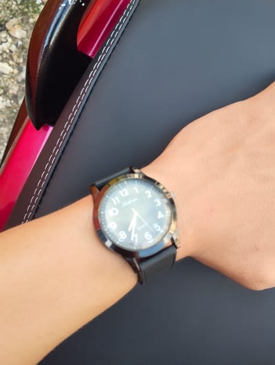The image displays a close-up shot of a person's wrist wearing a black-strapped watch. The watch has a round, dark face with white numbers and hands, indicating the time is approximately 10:40. The watch is positioned on a wrist with light-toned skin, which is resting on a textured black surface, possibly a seat or cushion. To the left of the wrist and black surface, a portion of a shiny, red object is visible, with white stitching along its edge where it meets the black surface. The background is out of focus, showing a rough, greyish texture that might be pavement or ground. No specific location within Pompanua, Indonesia is discernible from the image.